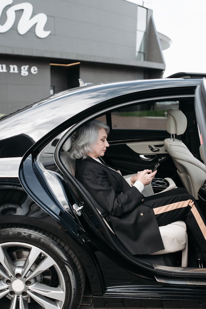 gallery-5 Senior woman in business attire sits in a luxury car, using a tablet outside a modern building.