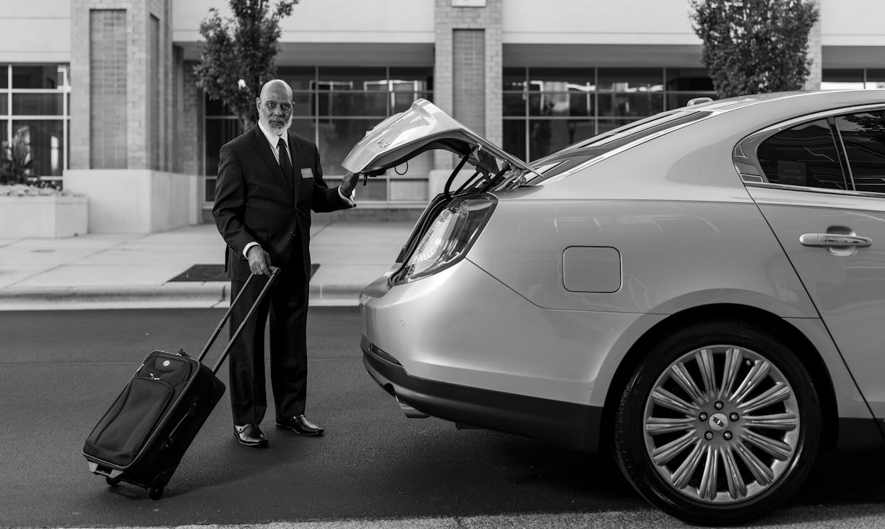 Chauffeur in suit loading luggage into luxury car trunk outside a modern building.