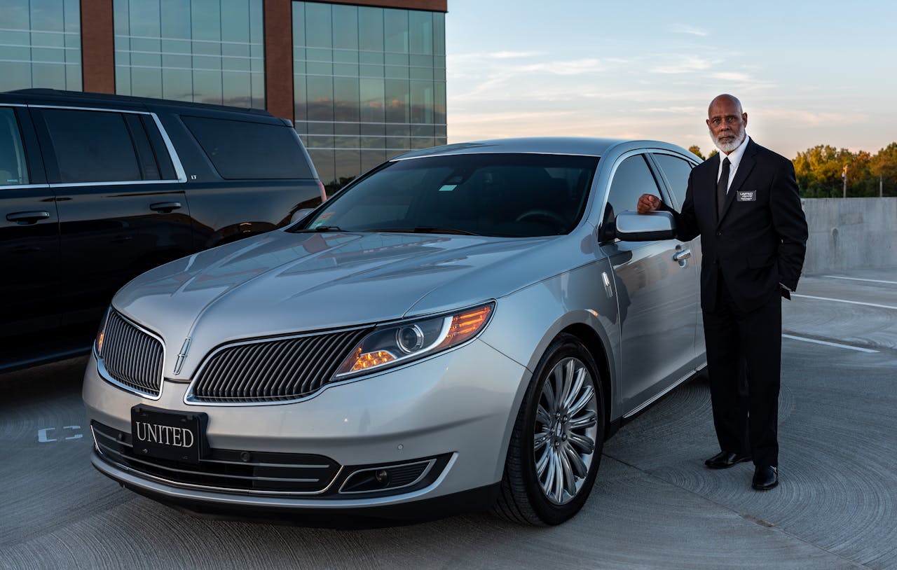 Professional chauffeur in a suit standing next to a luxury car in outdoor setting.