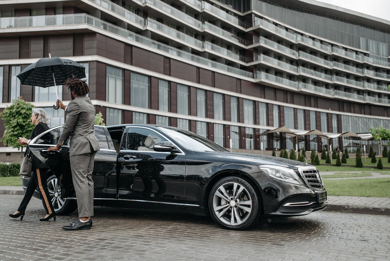 A business professional exits a luxury car while being shielded by an umbrella.